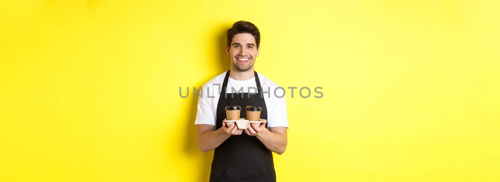 Handsome male barista serving takeaway coffee and smiling, bringing order, standing in black apron against yellow background by Benzoix