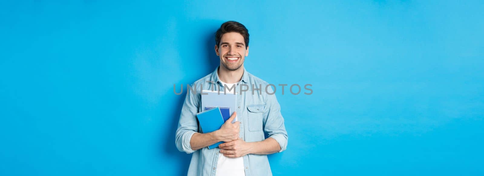 Young man holding notebooks and study material, smiling happy, standing over blue background by Benzoix