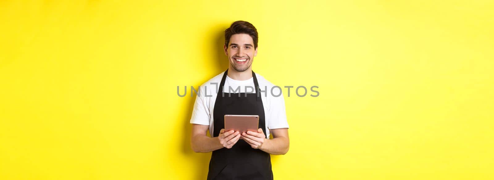 Waiter in black apron taking orders, holding digital tablet and smiling friendly, standing over yellow background by Benzoix