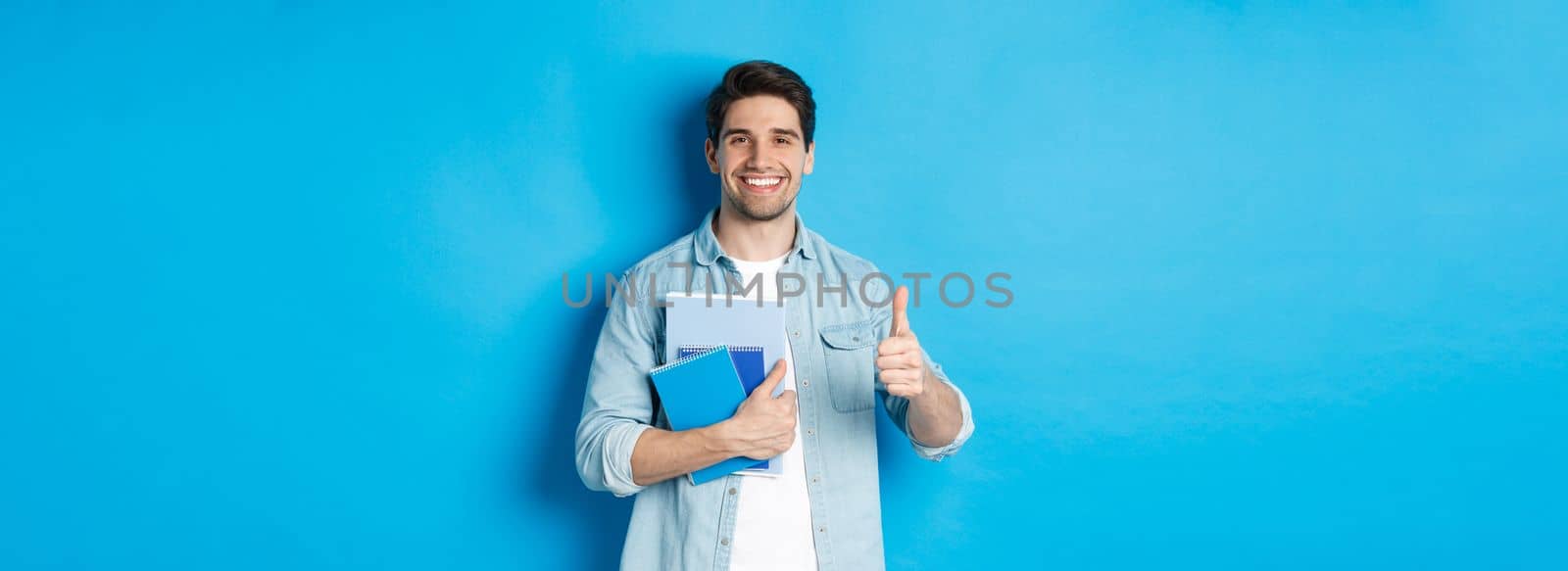 Young man student with notebooks, showing thumb up in approval, smiling satisfied, blue studio background by Benzoix