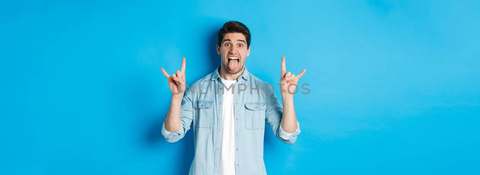 Cool guy having fun, enjoying concert, showing rock sign and tongue, standing over blue background.