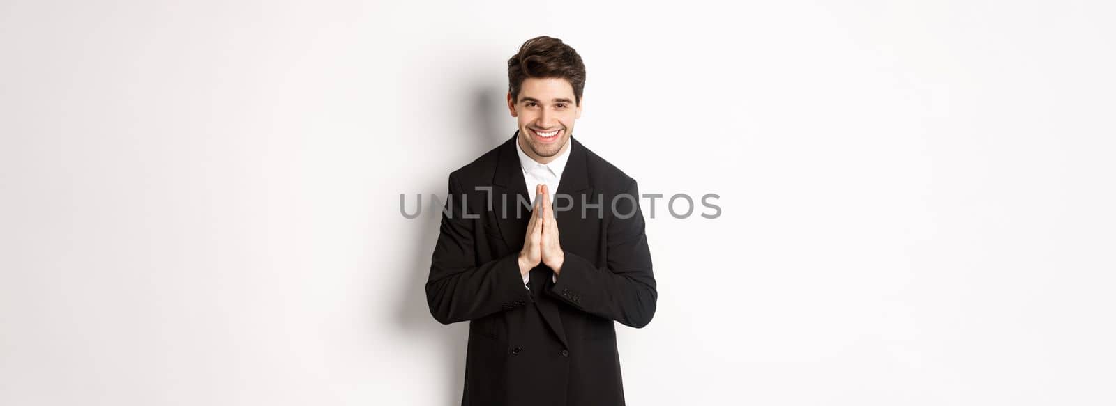 Portrait of handsome man in black suit, being grateful, saying thank you and bowing politely, smiling as holding hands together, expressing gratitude, standing over white background.