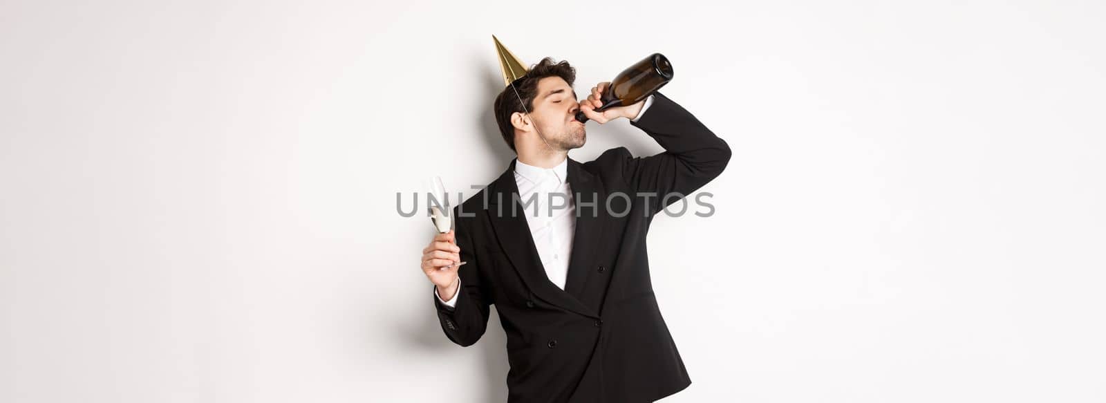 Image of handsome guy in trendy suit, drinking champagne from bottle and celebrating holiday, having a birthday party, getting drunk while standing over white background.
