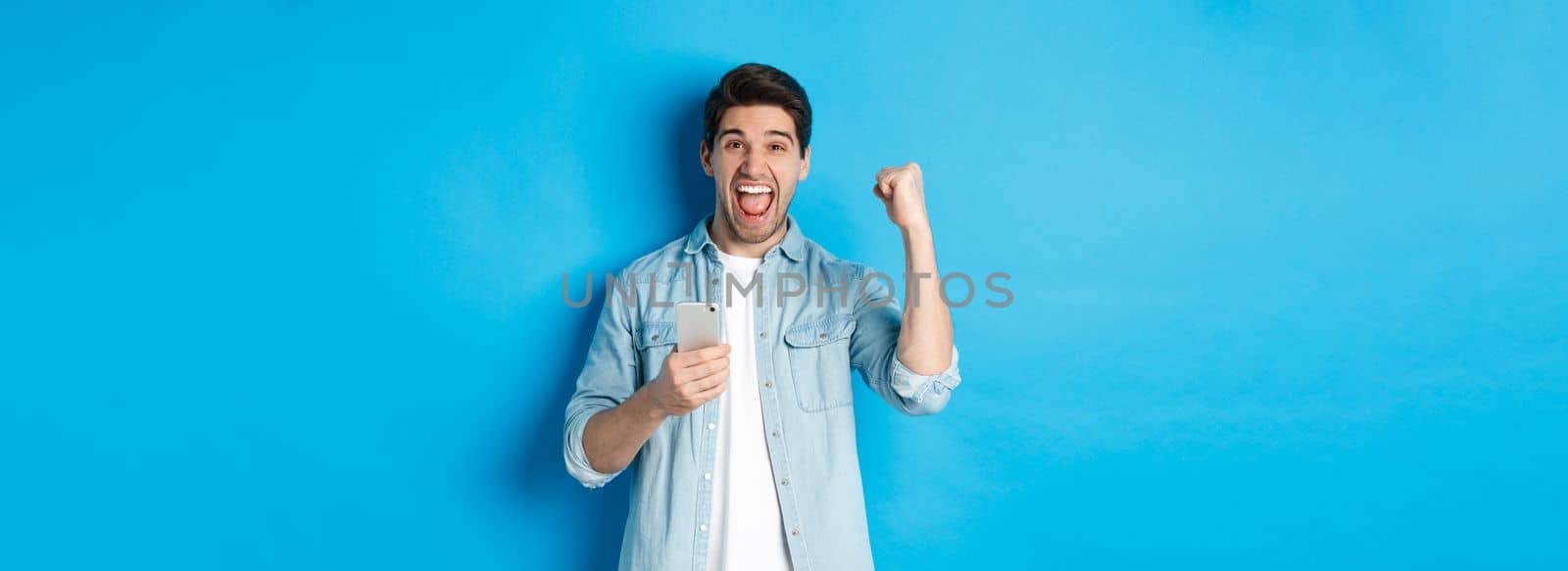 Concept of online shopping, applications and technology. Excited man shouting yes and making fist pump gesture after winning on smartphone, standing over blue background by Benzoix