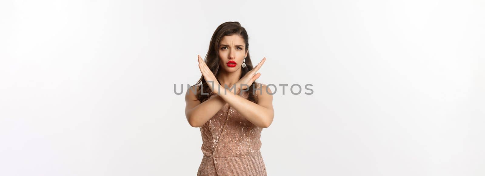 Displeased young woman in luxurious dress and red lipstick saying no, showing stop sign, cross gesture to refuse or prohibit, standing over white background.