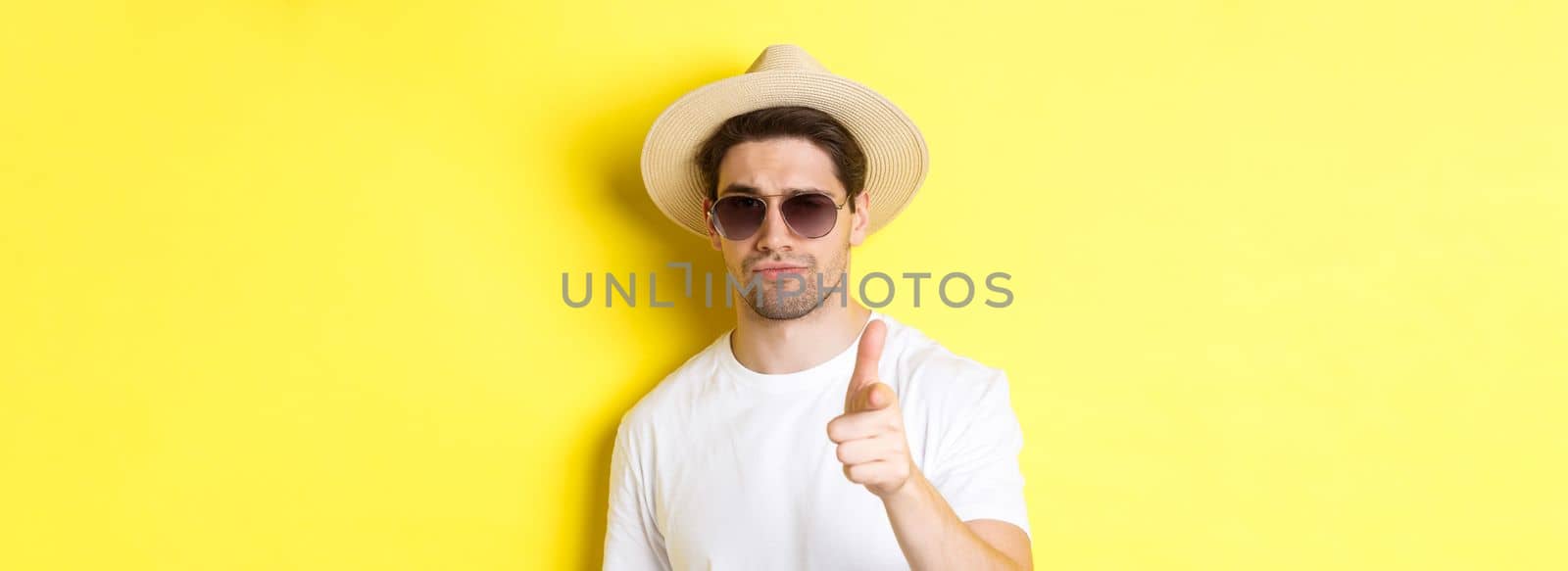 Concept of tourism and vacation. Close-up of cool guy in summer hat and sunglasses pointing finger at camera, standing over yellow background.