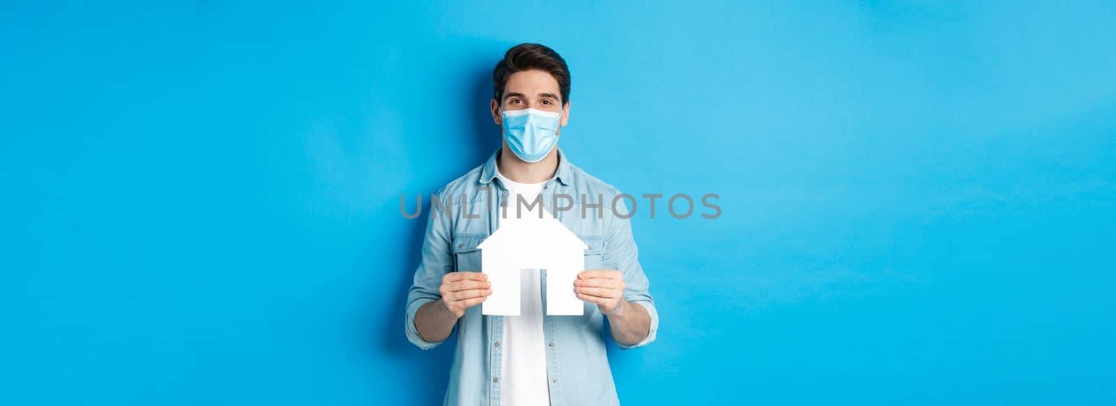 Concept of coronavirus, quarantine and social distancing. Young man searching apartment, showing house paper model, wearing medical mask, renting or buying propery, blue background.