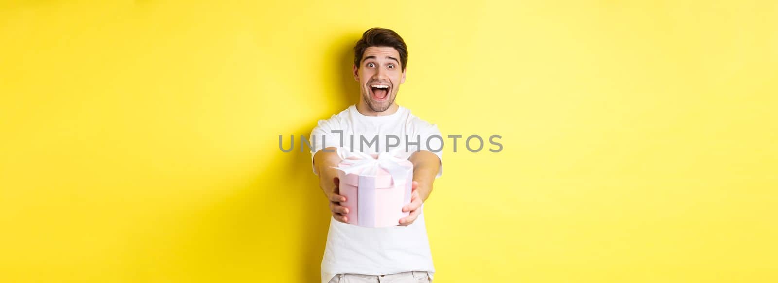 Concept of holidays and celebration. Happy man wishing happy new year, giving you gift, standing over yellow background.
