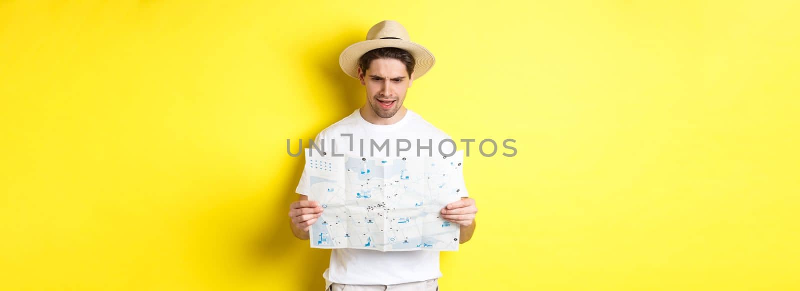Travelling, vacation and tourism concept. Man looking confused at map during trip, cant understand, standing over yellow background.