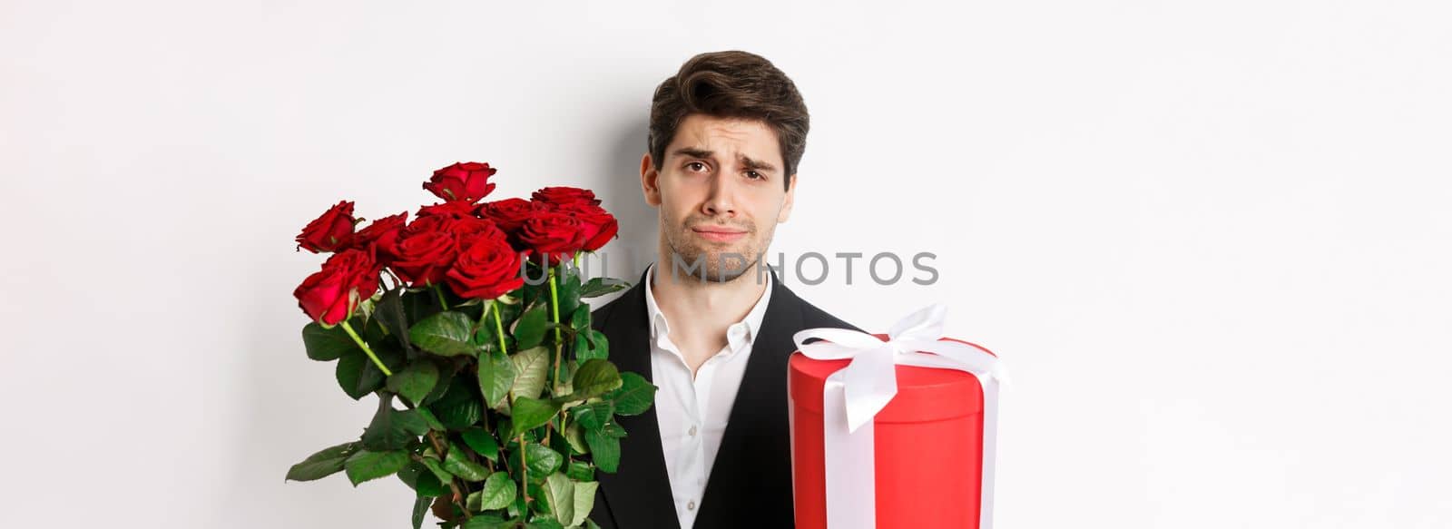 Close-up of sad man in suit, holding bouquet of red roses and a gift, standing upset against white background by Benzoix