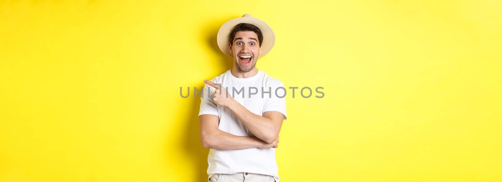 Happy male tourist in straw hat pointing finger right, showing promo offer on copy space, yellow background. Copy space