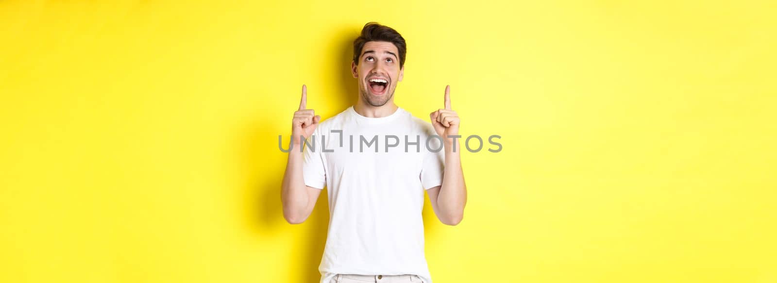 Cheerful young guy in white t-shirt reacting to promo offer, pointing and looking up with amazement, standing over yellow background.