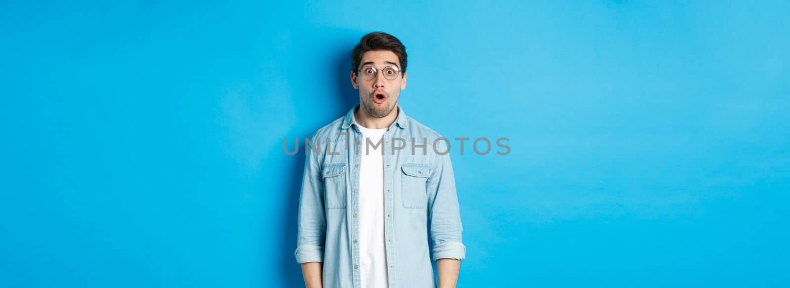 Surprised man in glasses looking at promo offer, saying wow amazed, standing over blue background.