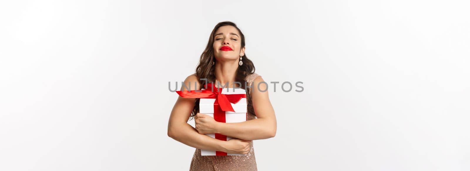 Holidays, celebration concept. Happy and thankful woman hugging Christmas gift, wearing luxury dress, receiving New Year present, standing over white background.