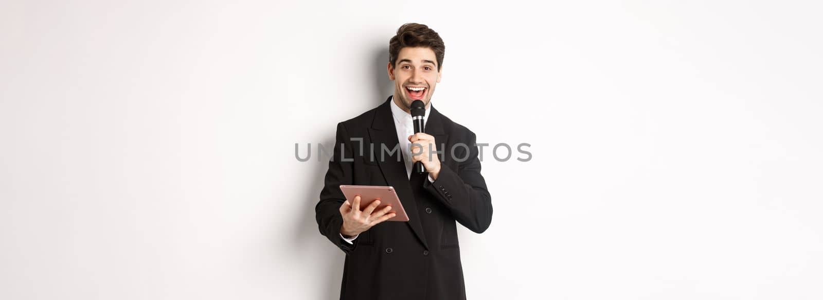 Image of handsome male performer, giving a speech, entertain people at party, holding microphone and digital tablet, standing over white background.