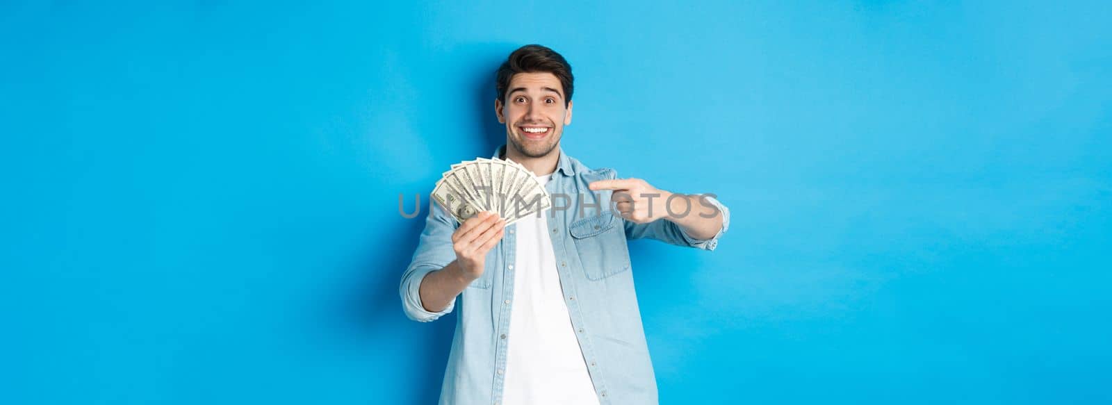 Surprised smiling man in casual clothes, pointing fingers at money, standing over blue background.