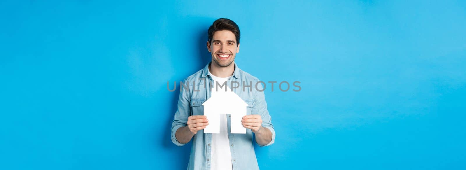 Insurance, mortgage and real estate concept. Smiling young man holding house model, searching apartment for rent, standing against blue background.