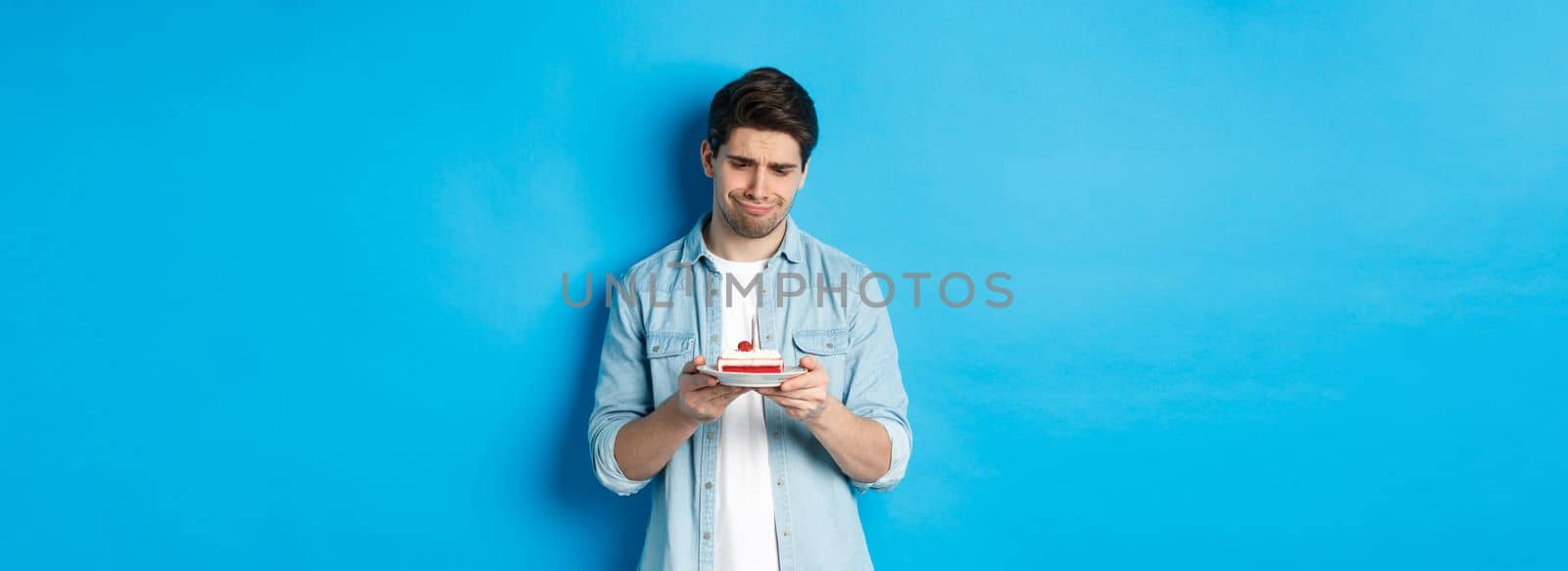 Holidays and celebration. Disappointed and sad man looking at birthday cake, grimacing upset, standing against blue background by Benzoix