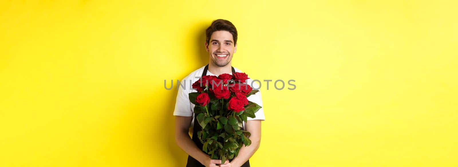 Smiling florist in black apron holding flowers, selling bouquet of roses, standing over yellow background.