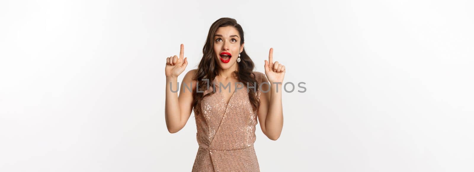 New Year, christmas and celebration concept. Excited beautiful woman showing advertisement, gasping amazed, wearing party dress and pointing up, white background.