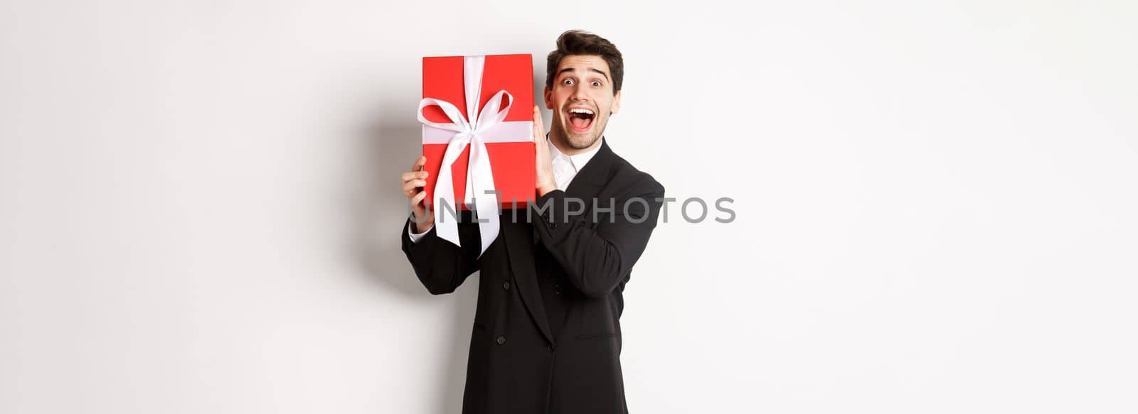 Image of happy good-looking man in black suit, holding box with christmas gift and smiling, standing against white background by Benzoix