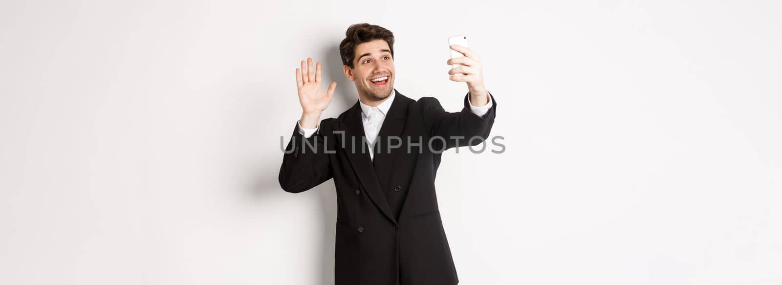 Image of handsome man in suit, having video call and waving hand at smartphone camera, recording video, greeting someone, standing against white background.