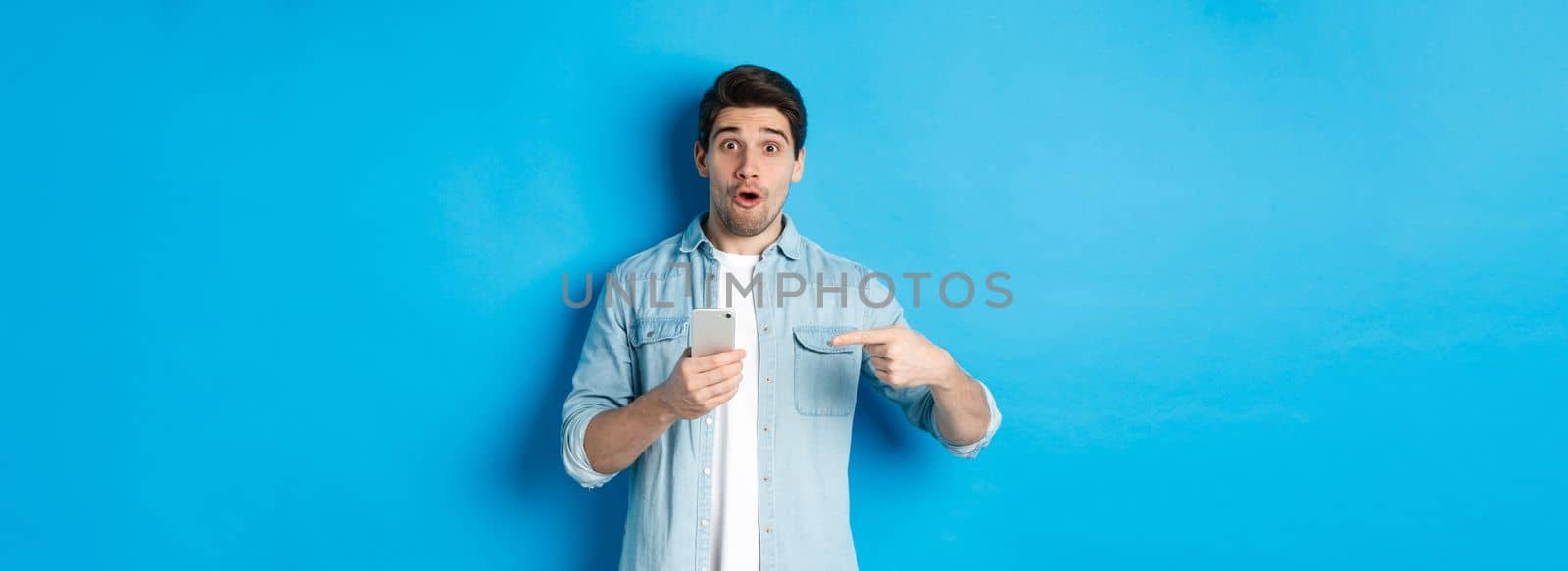 Concept of online shopping, applications and technology. Impressed man pointing finger at mobile phone and looking amazed, recommending app, standing over blue background.