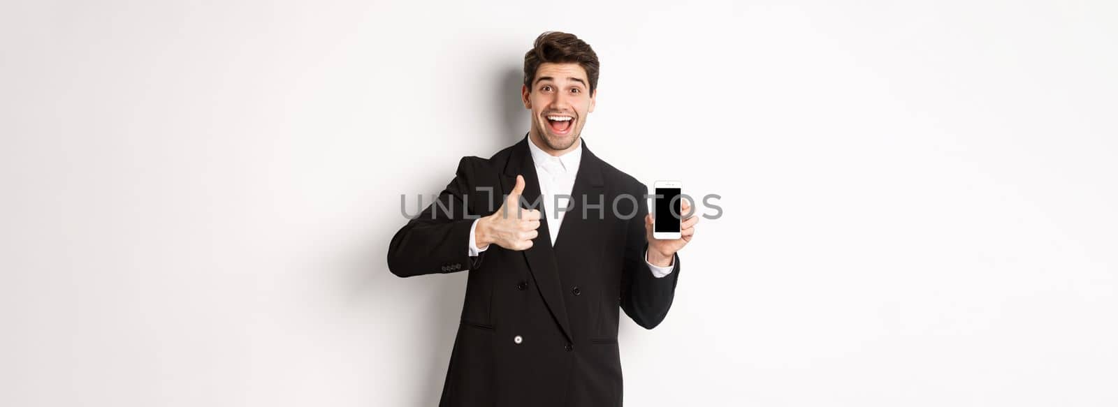 Image of handsome male entrepreneur in black suit, recommending app or online shop, showing thumbs-up and smartphone screen, standing over white background.