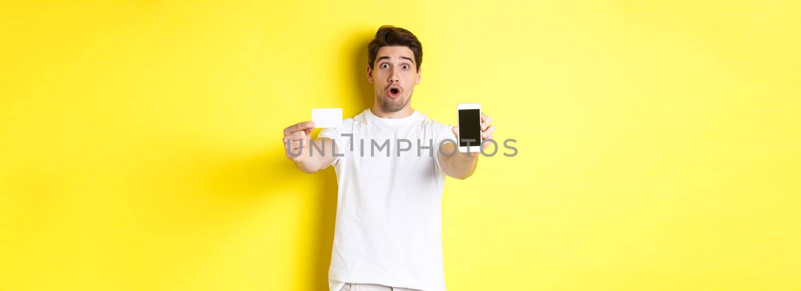 Handsome caucasian guy showing smartphone screen and credit card, concept of mobile banking and online shopping, yellow background.
