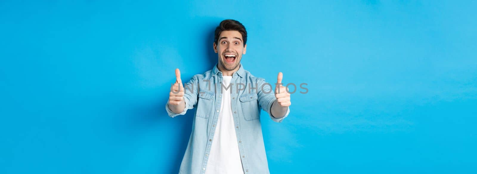 Smiling confident man showing thumbs up with excited face, like something awesome, approving product, standing against blue background.