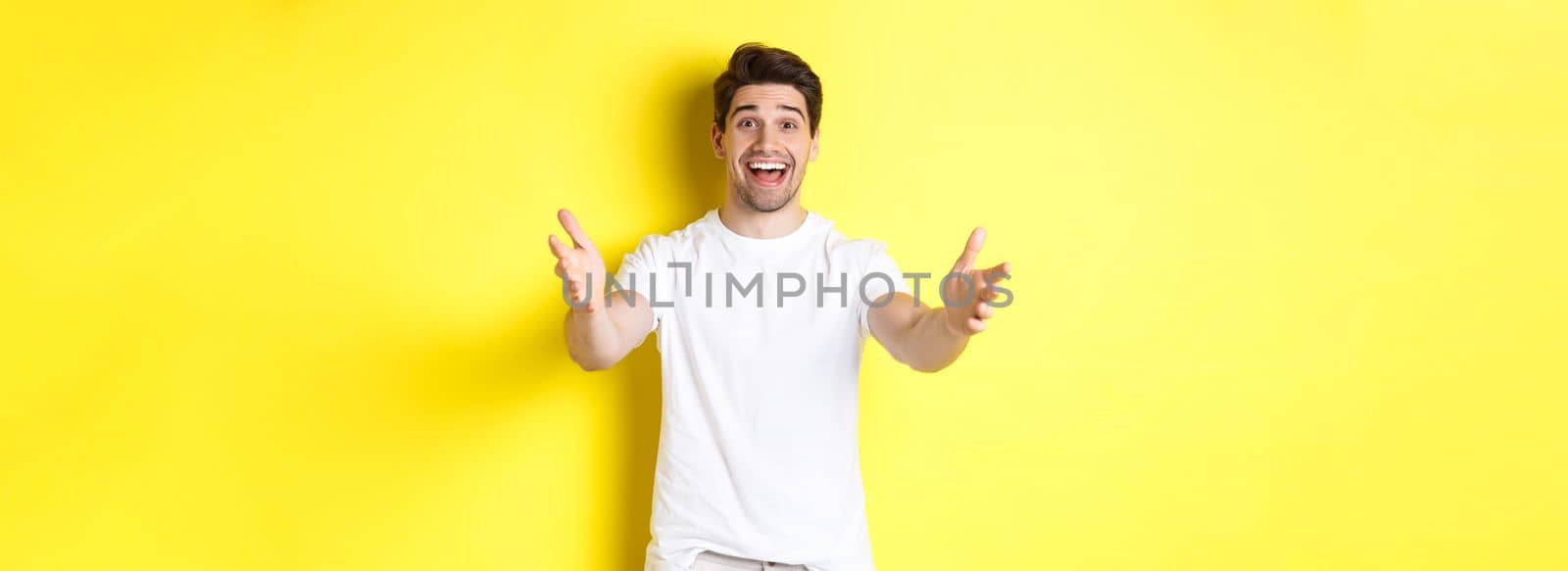 Excited handsome guy stretching hands forward, reaching for hug, receiving gift, standing over yellow background by Benzoix