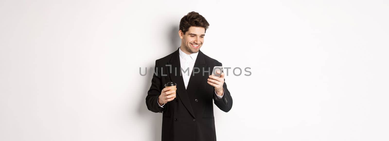 Portrait of handsome, confident businessman in black suit, drinking coffee and using mobile phone, smiling pleased, standing over white background by Benzoix