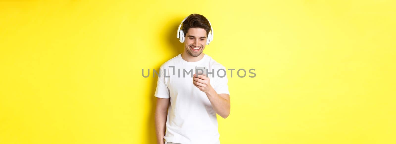 Young man in headphones reading message on smartphone, smiling, listening music in earphones and picking song in app, standing over yellow background by Benzoix