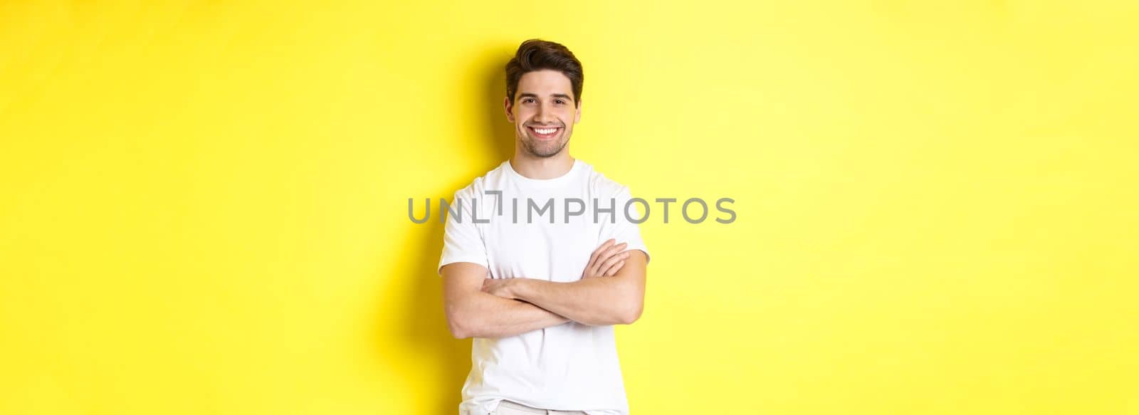 Image of confident caucasian man smiling pleased, holding hands crossed on chest and looking satisfied, standing over yellow background by Benzoix