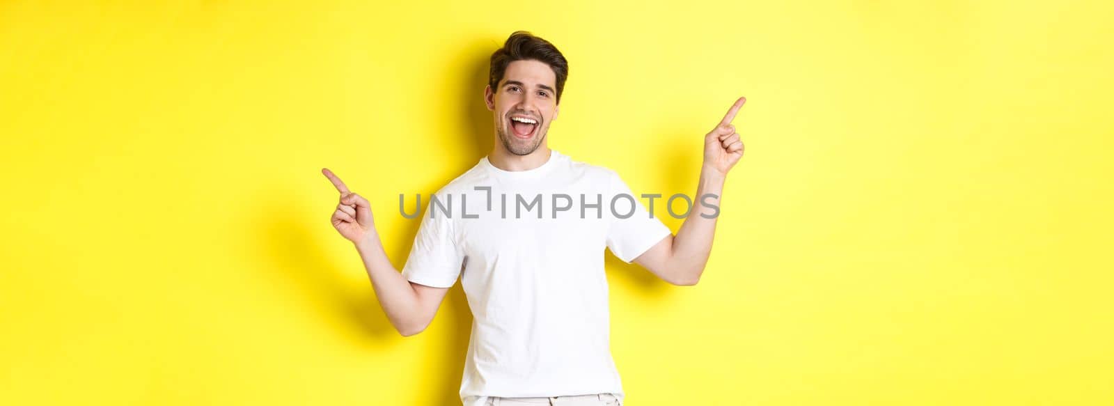 Happy stylish guy showing two variants, pointing fingers sideways at left and right promo, standing over yellow background.