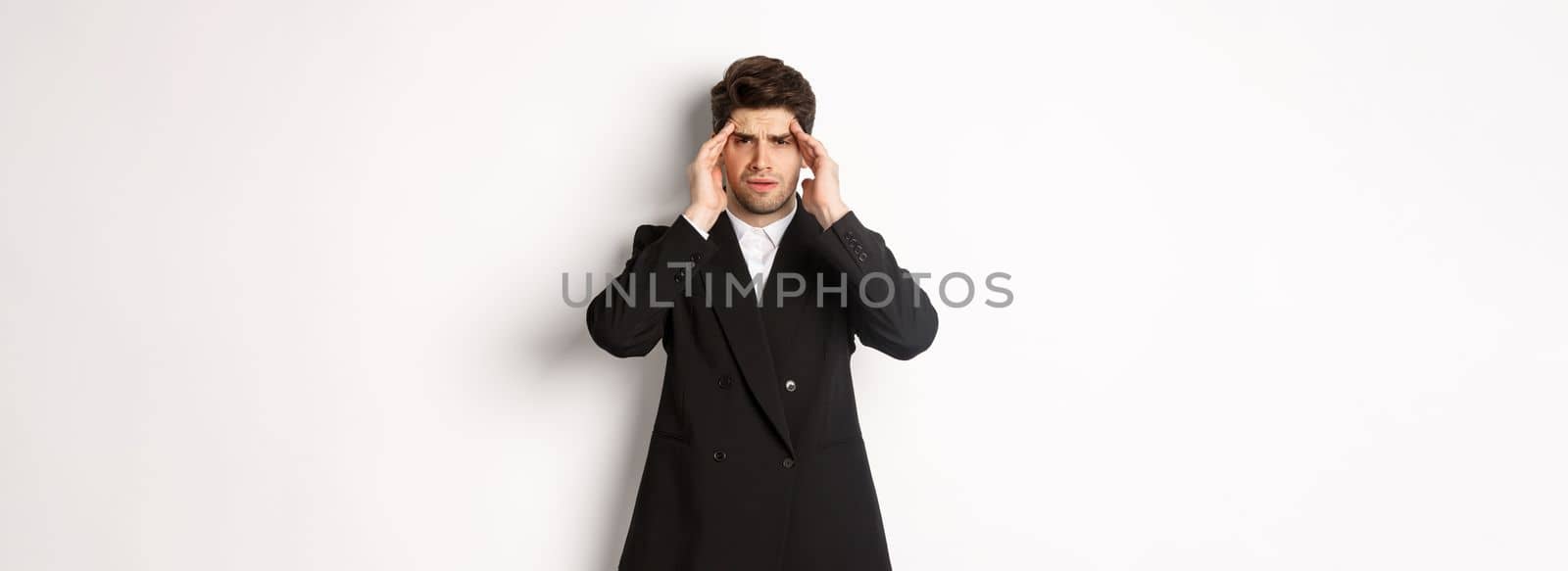 Image of businessman in black suit, touching head and looking dizzy, feeling painful headache, standing over white background.