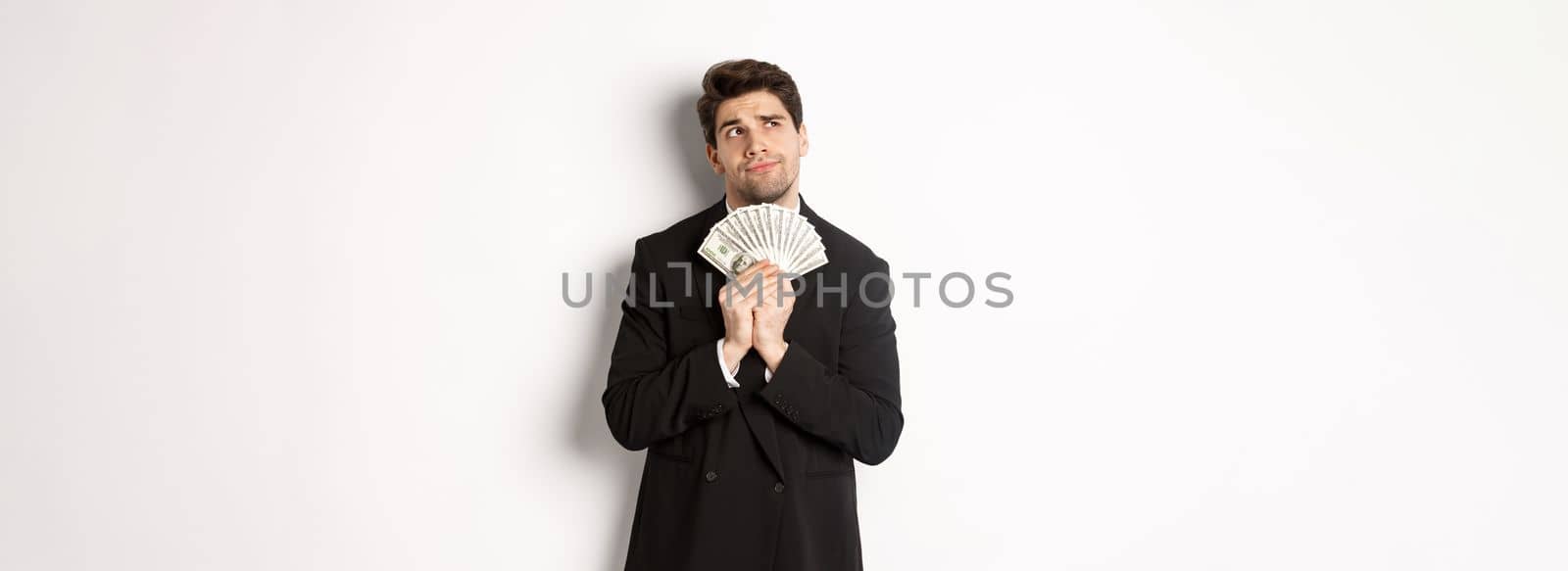 Image of handsome businessman in black suit, holding money and thinking about investment, making shopping plans, standing over white background by Benzoix
