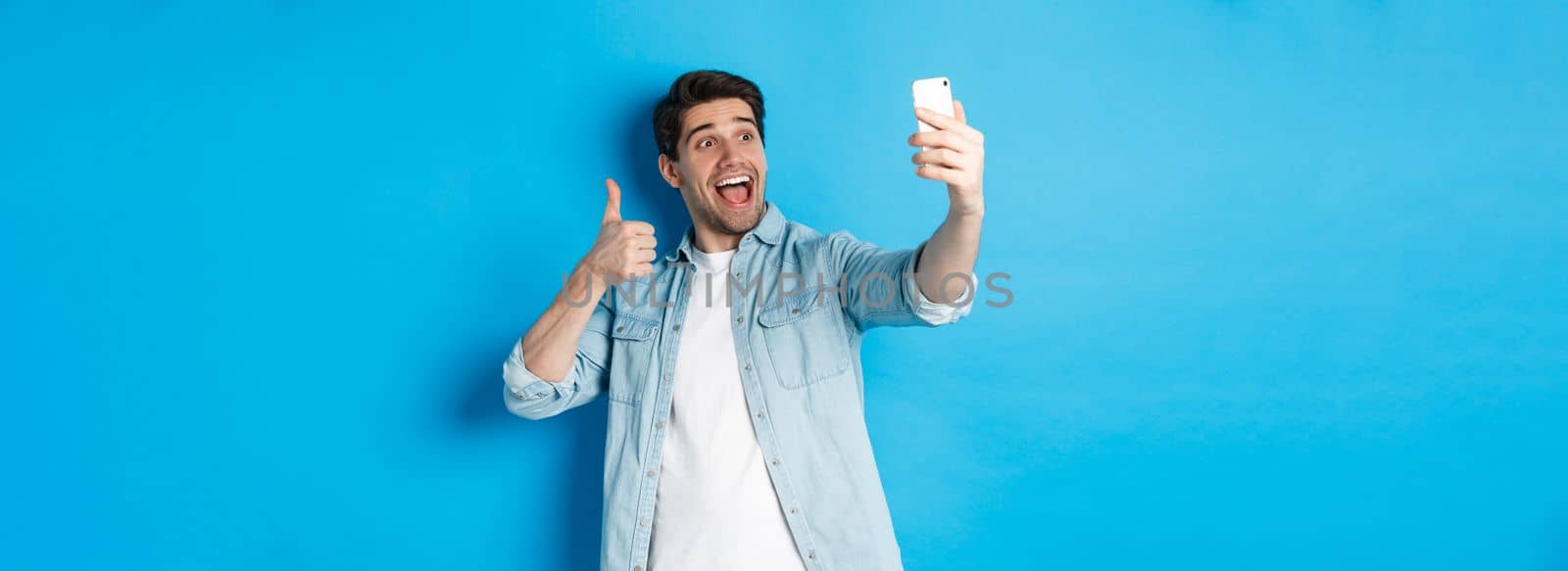 Happy man taking selfie and showing thumb up in approval on blue background, holding mobile phone.