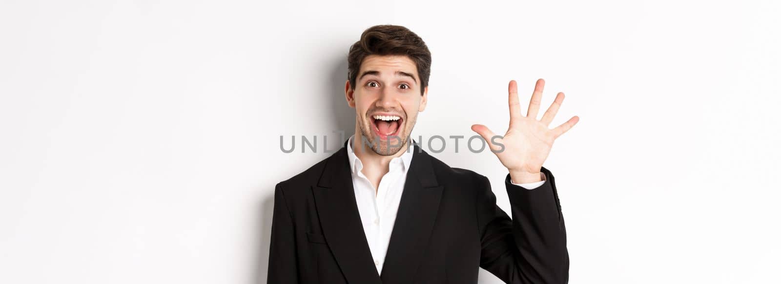 Close-up of handsome businessman in black suit, smiling amazed, showing number five, standing over white background.