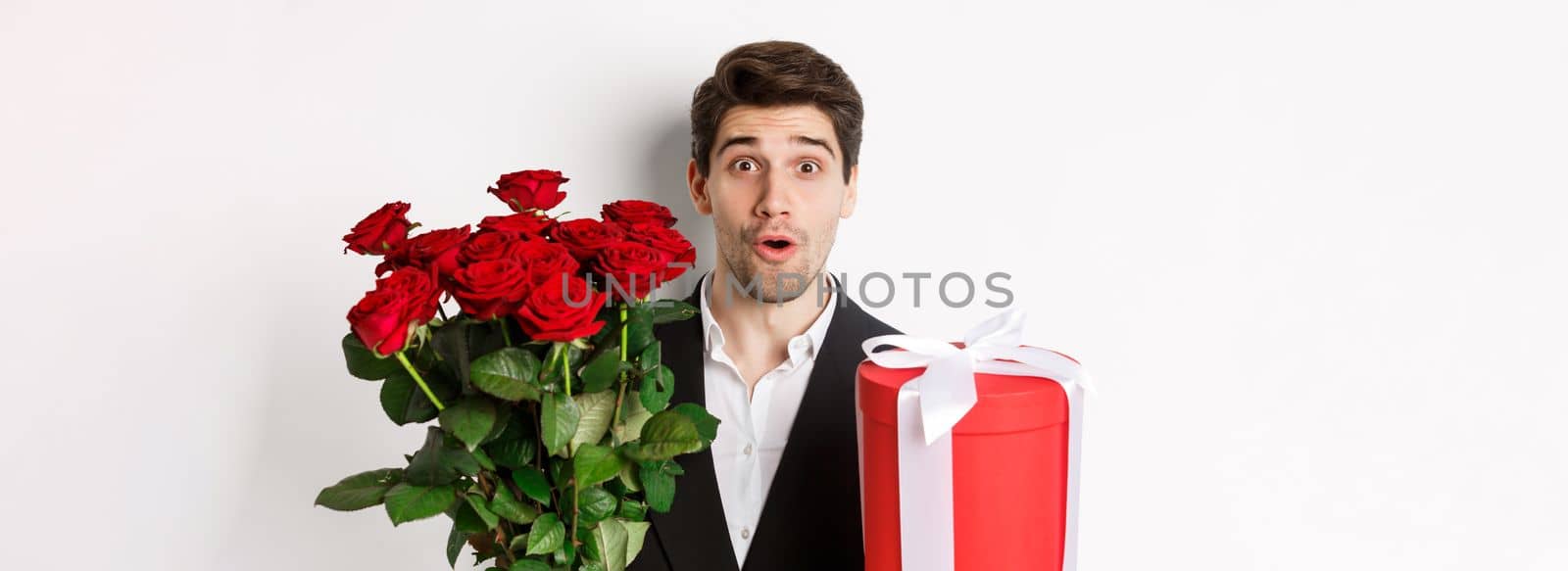 Close-up of attractive man in suit looking surprised, holding gift box and bouquet of roses, giving present for holiday, standing against white background by Benzoix