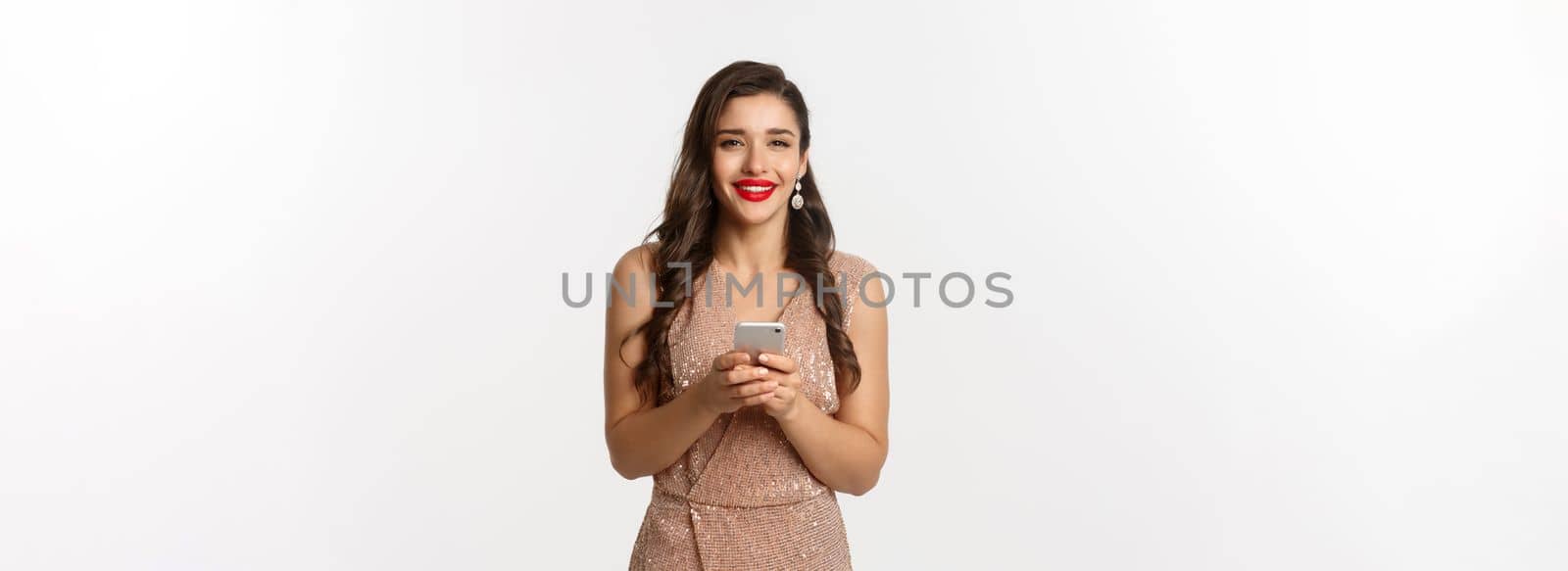 Christmas party and celebration concept. Happy young woman in elegant dress using mobile phone and smiling, shopping online, standing over white background by Benzoix