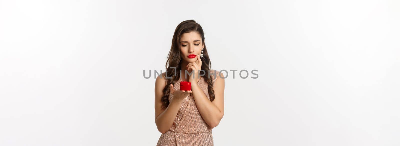 Image of beautiful woman thinking about marriage proposal, looking at engagement ring box and thinking, standing over white background in glamour dress.