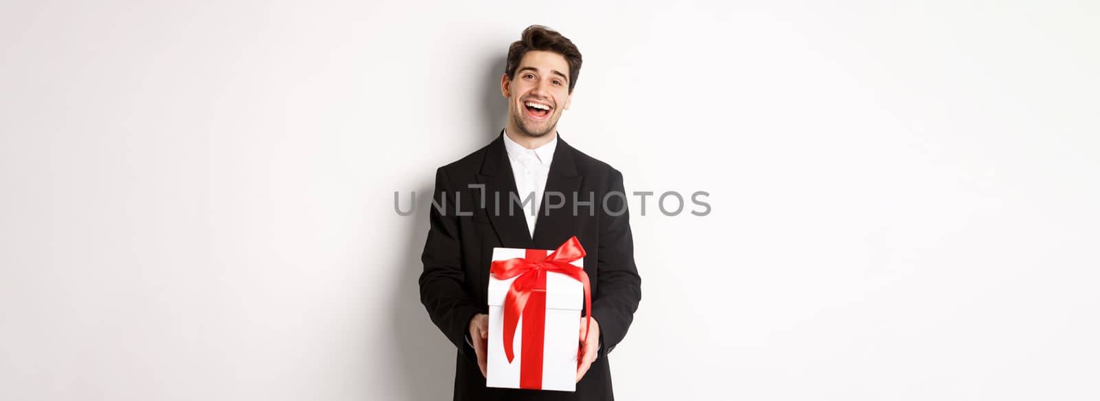 Concept of christmas holidays, celebration and lifestyle. Joyful handsome man in black suit, holding xmas gift and smiling, standing against white background.