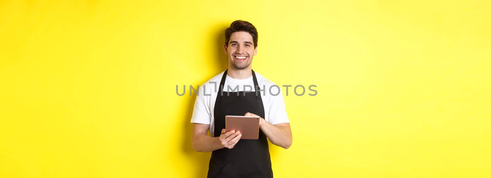 Handsome waiter taking orders, holding digital tablet and smiling, wearing black apron uniform, standing over yellow background by Benzoix