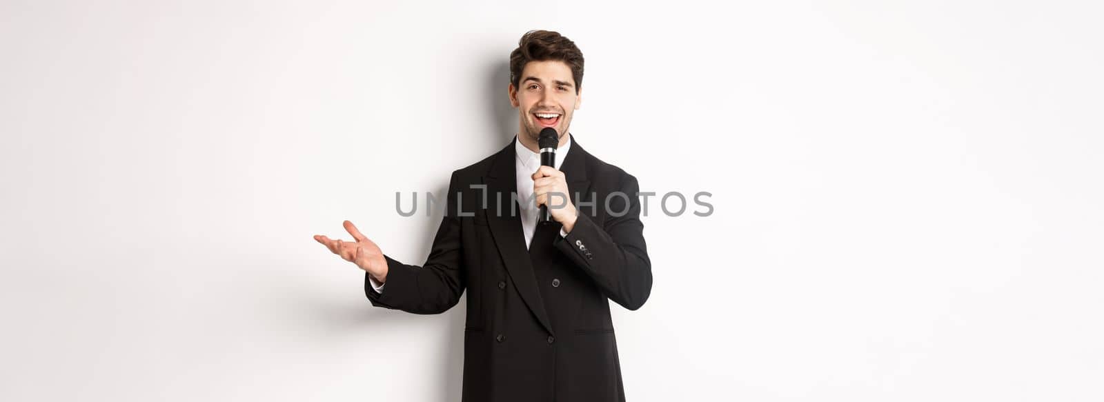 Portrait of handsome man in black suit singing a song, holding microphone and giving speech, standing against white background.