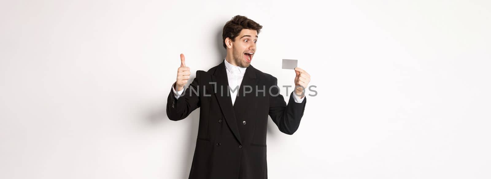 Image of excited handsome businessman, showing credit card and thumb-up, standing against white background by Benzoix