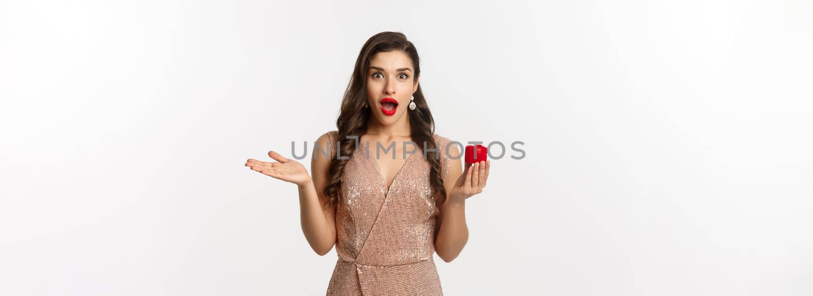 Image of amazed girlfriend holding engagement ring box, looking surprised, receive marriage proposal, wearing elegant dress, white background.