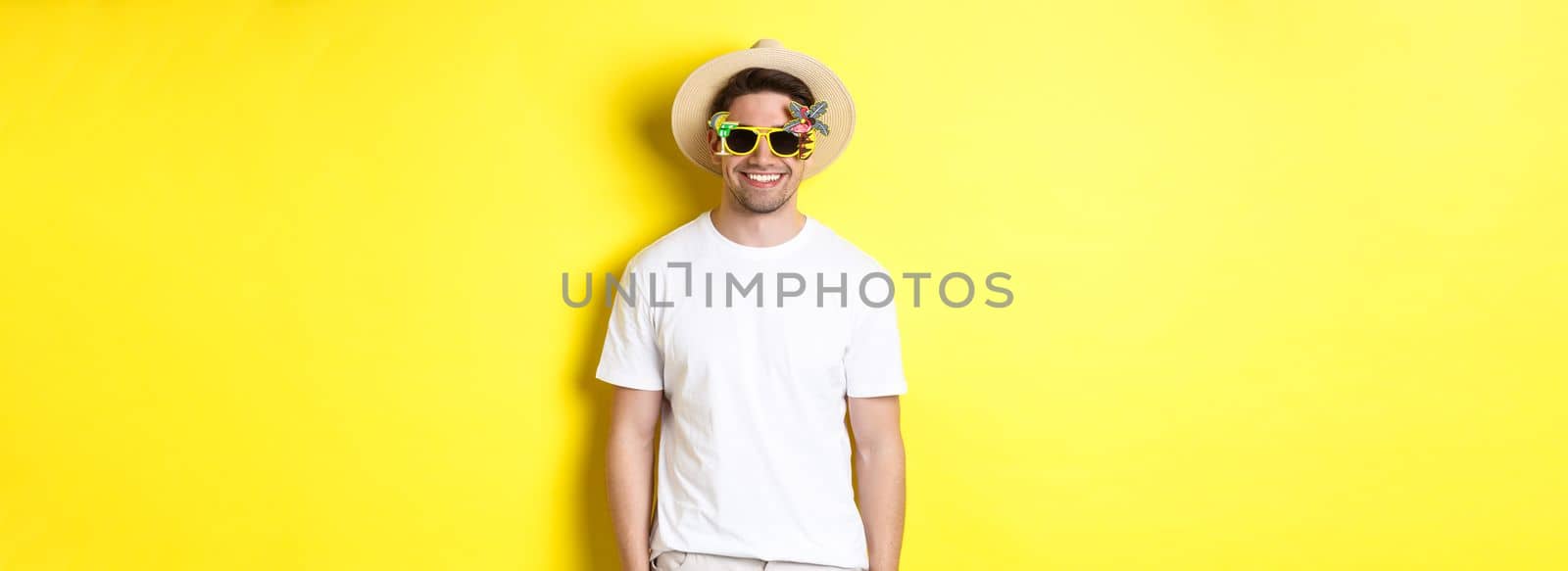 Concept of tourism and vacation. Relaxed smiling man enjoying supper trip, wearing sunglasses and straw hat, yellow background.