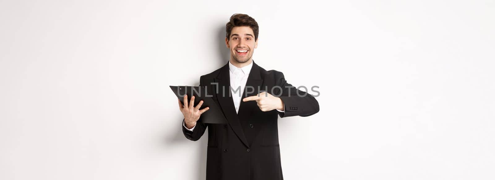 Image of handsome smiling businessman in black suit, pointing finger at clipboard with documents, standing against white background.