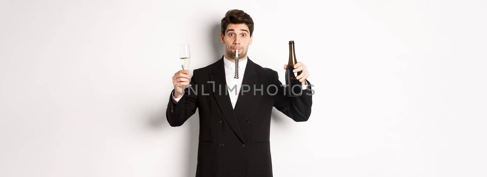 Concept of holidays, party and celebration. Portrait of handsome guy in black suit, raising bottle of champagne and glass, blowing a party whistle, having a birthday, standing over white background by Benzoix